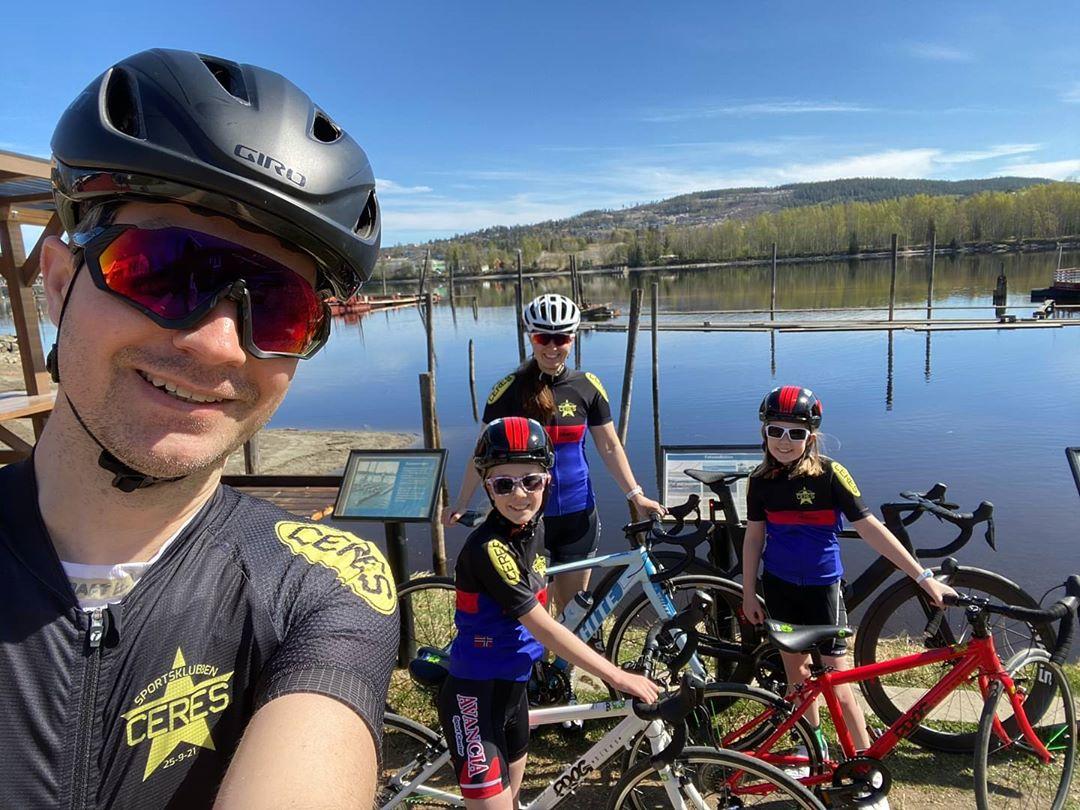 Dad taking a selfie photo with daughters and wife in cycling kit by a lake all on their road bikes